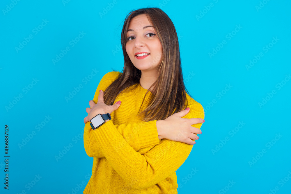 Charming pleased Young arab woman wearing knitted sweater over blue backgtound embraces own body, pleasantly feels comfortable poses. Tenderness and self esteem concept Charming pleased Young arab woman wearing knitted sweater over blue backgtound embraces own body, pleasantly feels comfortable poses. Tenderness and self esteem concept