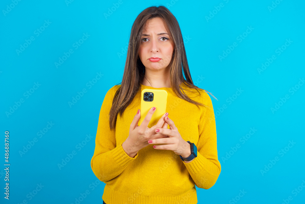 Upset dissatisfied Young arab woman wearing knitted sweater over blue backgtound uses mobile software application and surrfs information in internet, holds modern mobile hand Upset dissatisfied Young arab woman wearing knitted sweater over blue backgtound uses mobile software application and surrfs information in internet, holds modern mobile hand