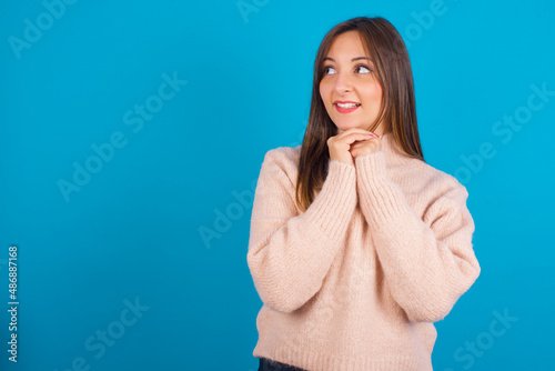 Happy Young arab woman wearing knitted sweater over blue backgtound anticipates something awesome happen, looks happily aside, keeps hands together near face, has glad expression. © Roquillo