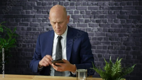 Mature businessman in suit hangs up phone in office. The bald senior man is talking on his cellphone before ending the call.