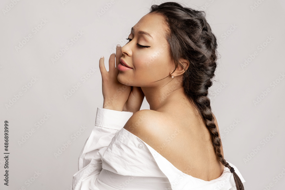 Sensual pretty mixed race young woman with bare shoulder, long pigtail, perfect make up posing on gray studio background. Skincare, make-up cosmetics advertising. Natural womens beauty concept. Sensual pretty mixed race young woman with bare shoulder, long pigtail, perfect make up posing on gray studio background. Skincare, make-up cosmetics advertising. Natural womens beauty concept.
