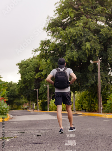 Man with backpack wearing cap, shorts and t-shirts walking in the middle of the road. Backpacker hiking in Caribbean country.