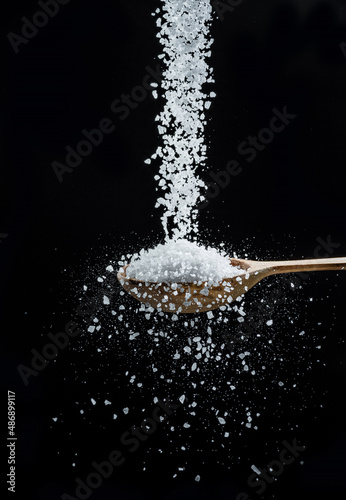 Edible salt crystals falling down into the wooden spoon at black background.