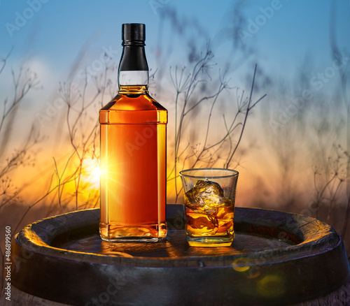 Whisky bottle and glass of whisky on old wooden cask. Beautiful sunset at the background.