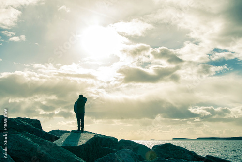 Ein Mann steht auf eine Felsgruppe und schaut gegen die Sonne übers Meer