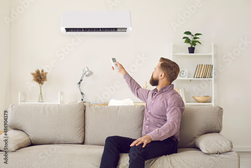 Photos Male renter relax on comfortable sofa hold remote controller turn conditioner for fresh ventilated air at home