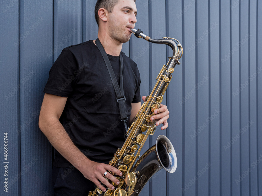 Fototapeta premium Young street musician playing saxophone near the big blue wall