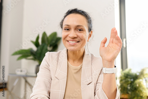 Portrait of smiling and friendly multi ethnic female freelancer looking at the camera and greeting. Cheerful woman in smart casual wear waving, while working remotely
