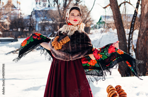 Outdoors lifestyle portrait of pretty young woman in a traditional Russian headscarf with bagels on winter background. Smiling and dancing on the folk fest. Shrovetide. Wearing a Russian folk clothes.