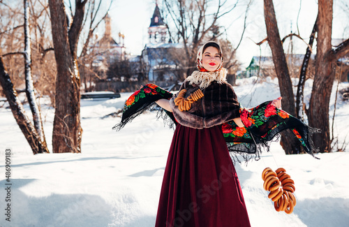 Outdoors lifestyle portrait of pretty young woman in a traditional Russian headscarf with bagels on winter background. Smiling and dancing on the folk fest. Shrovetide. Wearing a Russian folk clothes.