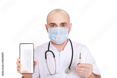 Middle-aged doctor in white robe with stethoscope on his neck on a white background holding a phone with a positive test and a syringe with the inscription covid