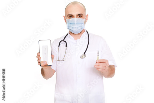 Middle-aged doctor in white robe with stethoscope on his neck on a white background holding a phone with a positive test and a syringe with the inscription covid