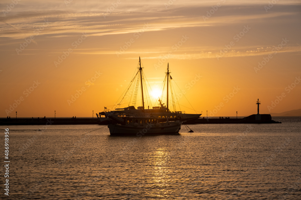 Sunset over Mykonos island, Cyclades, Greece. Moored ship at port, beacon orange sky sparkle sea.