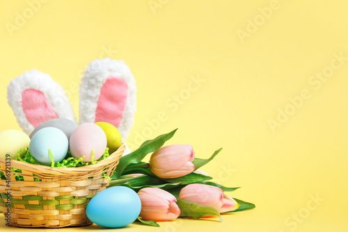 Easter composition. Eggs in a basket tulips rabbit ears on a yellow background.