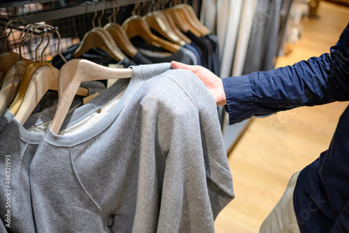Male hand customer choosing lightweight sweater hanging on rack in clothes shop. Fashion product collection in clothing store for selling. Textile industry and business concept