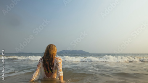 Crazy Woman in a White Tunic on the Beach, near the Stormy Sea. Cheerful Funny Charming Blonde with Wet Curly Hair, in Sunglasses. Smiling Happy Girl Tourist Lies in the Water, has Fun. Back View © ivandanru