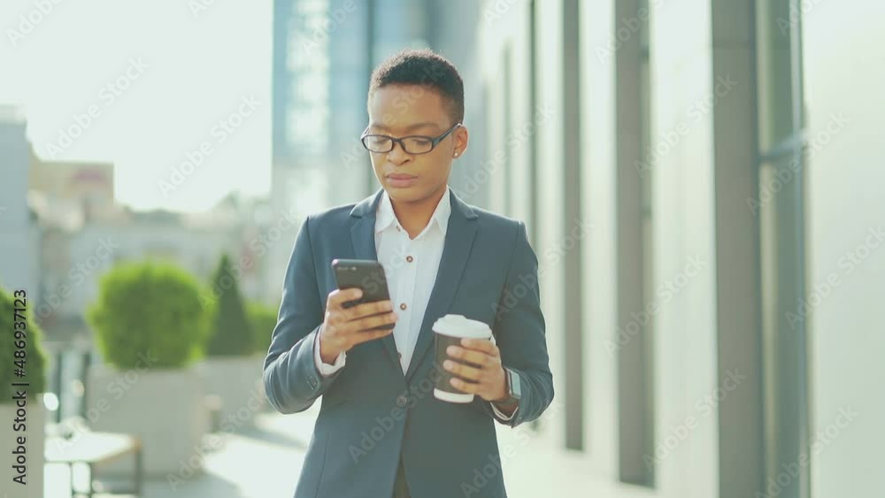 business black woman walking on city street downtown with mobile phone and cup coffee. African American female employee entrepreneur uses a smartphone on urban background. confident office worker business black woman walking on city street downtown with mobile phone and cup coffee. African American female employee entrepreneur uses a smartphone on urban background. confident office worker