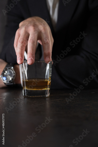 Hand of a man in a white shirt and dark suit holds a glass of whiskey on the bar