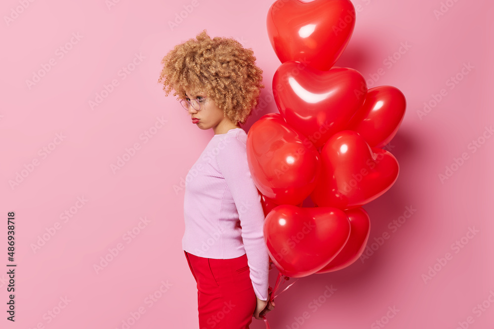Offended young woman with curly hair stands sideways at camera holds bunch of inflated balloons behind back feels stressed and lonely isolated over pink background. Sad Valentines Day concept Offended young woman with curly hair stands sideways at camera holds bunch of inflated balloons behind back feels stressed and lonely isolated over pink background. Sad Valentines Day concept