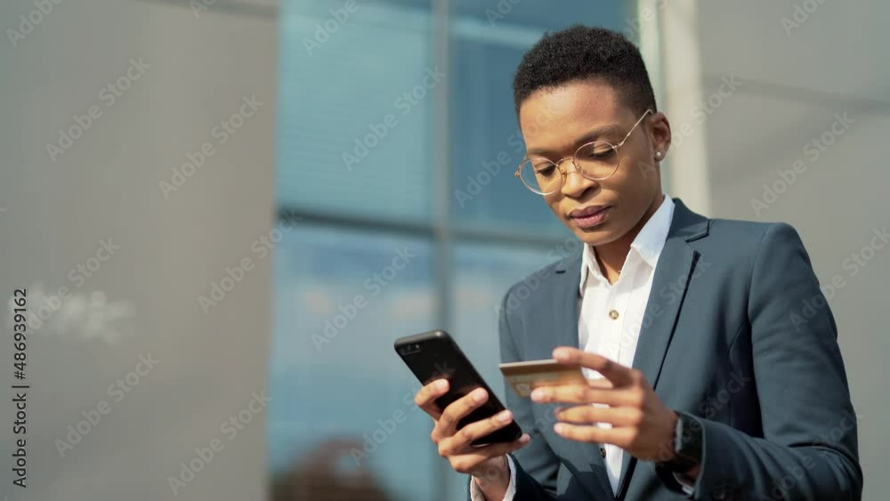 African American business woman shopping or making payment using mobile phone smartphone and credit card. female office worker employee buyer on outside sits on a bench, enters data Outdoor urban park