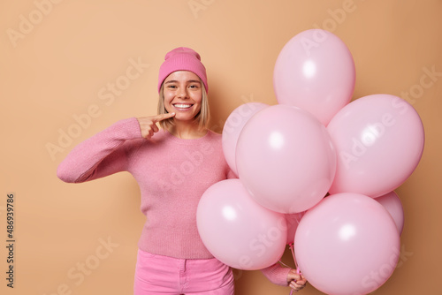 Indoor shot of happy young woman spends free time on hen party points at her toothy smile holds big bunch of helium balloons wear spink hat jumper and trousers isolated over brown background.