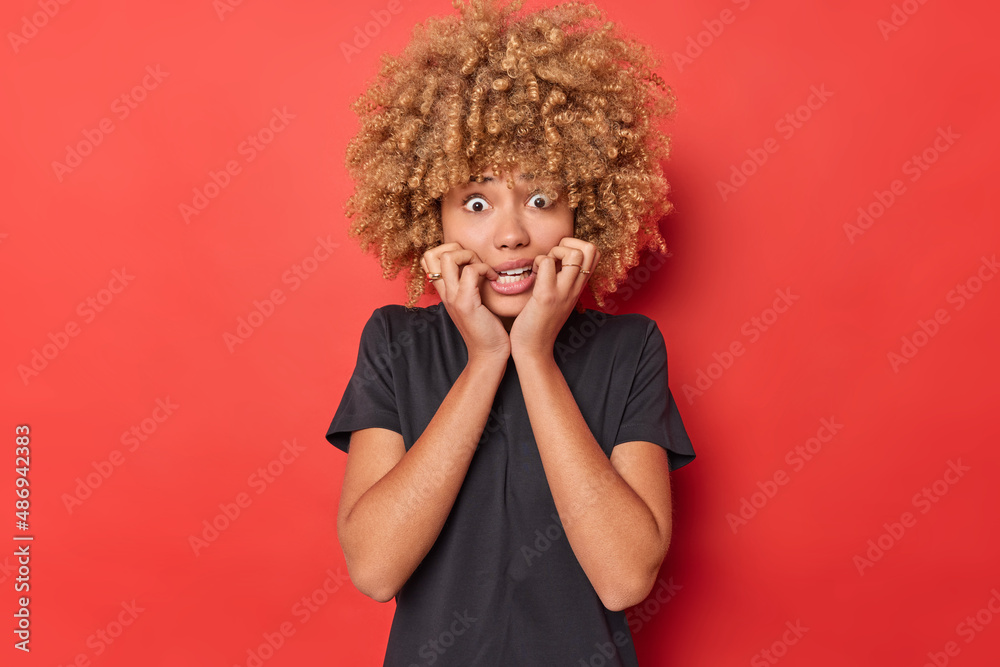 Anxious scared woman bites finger nails stans worried being insecure of something wears casual black t shirt isolated over vivid red background. Terrified frightened girl sees horrible scene in front