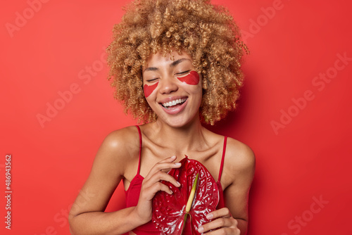 Positive carefree woman with curly bushy hair applies hydrogel patches under eyes to remove puffiness smiles broadly undergoes beauty treatments isolated over vivid red background. Beauty time