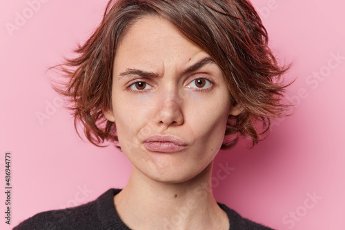 Headshot of serious beautiful young woman looks skeptical raises eyebrows purses lips considers something tries to make decision isolated over pink background. Human face expressions concept
