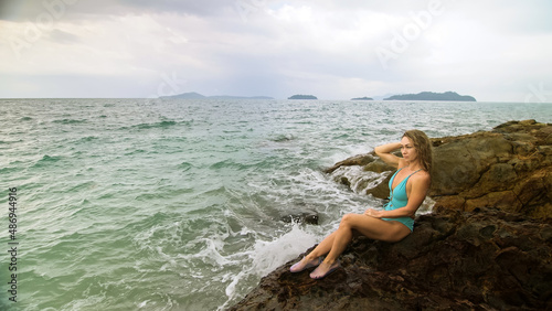 Handsome long-haired woman chill relax resting on rock of sea reef stone, stormy cloudy ocean. Woman in blue swimsuit dress tunic. Concept rest tropical resort coastline traveling outdoor tourism