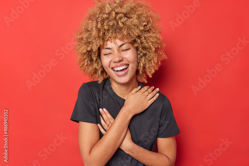 Happy carefree woman keeps arms crossed over chest laughs sincerely smiles broadly keeps eyes closed wears casual black t shirt poses against vivid red background. Positive emotions concept.