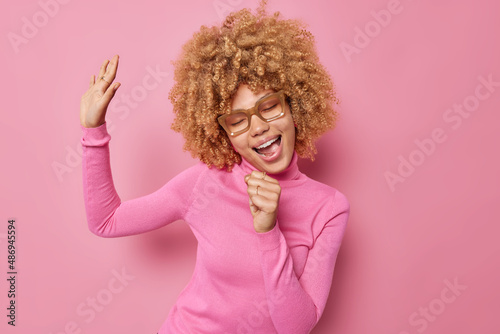 Horizontal shot of happy carefree woman keeps hand near mouth sings favorite song and dances has upbeat mood during spare time wears spectacles and casual turtleneck isolated on pink studio wall