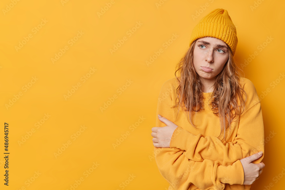 Frustrated young European woman looks sadly away feels offended wears casual jumper and hat feels displeased wears casual jumper and hat isolated over yellow background blank space for your promo