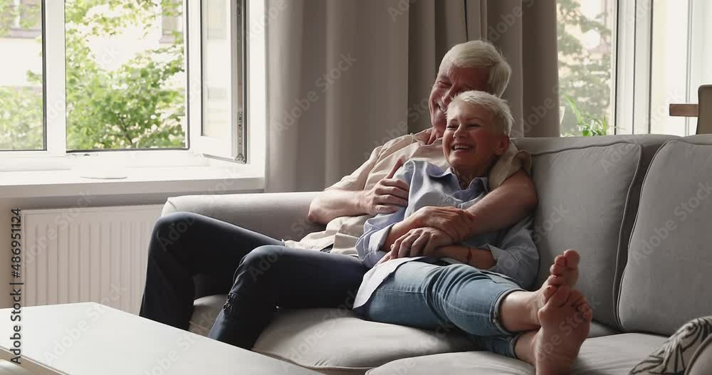 Positive smiling older grey-haired couple in love hugging daydreaming relaxing on cozy sofa enjoy pleasant conversation spend time at modern home. Happy marriage, harmony, romantic relations concept