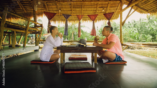 Attractive young man and woman eat and drink tasty beverages spending time in traditional local floating cafe on water. Loving happy couple having breakfast outdoors, feed each other. Tropical view