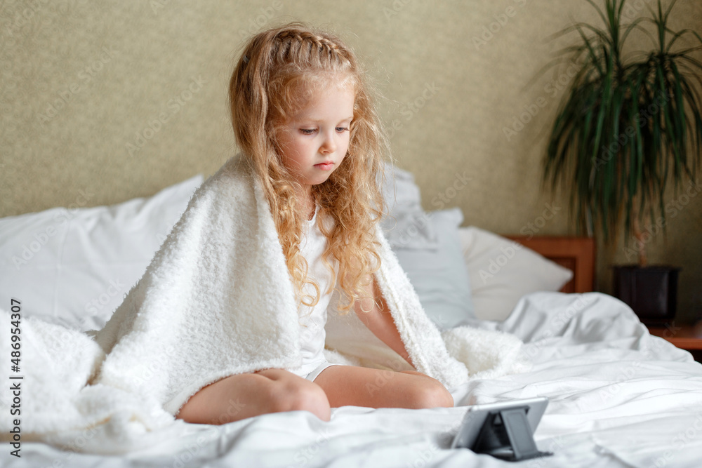Borring Preschooler girl watching video cartoons on smartphone at home. Beautiful caucasian kid bored sitting on bed covered with blanket in pajamas use phone. Unhappy child spend leisure time at home