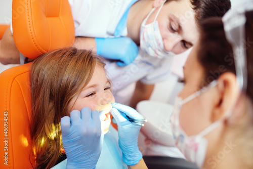 young dentists  a man and a woman  examine the teeth of a child s patient - a little pretty girl who is sitting in an orange dental chair.