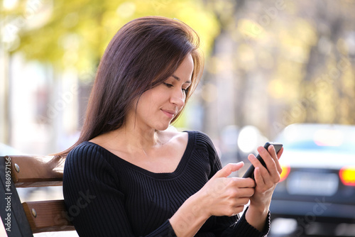 Young pretty woman messaging on mobile phone on warm summer day sitting on a city street bench outdoors