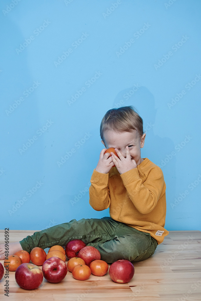 Fototapeta premium a boy of 2 years sits on a wooden table and plays with apples and tangerines, he closes his eyes and plays a game.