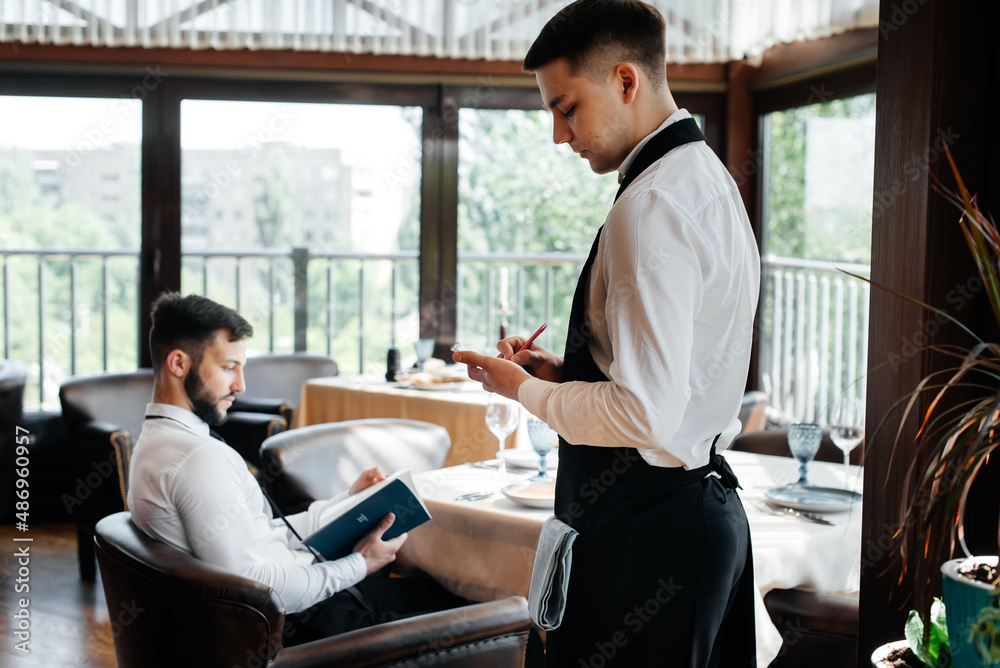 A young businessman in a fine restaurant examines the menu and makes an order to a young waiter in a stylish apron. Customer service. Table service in the restaurant. A young businessman in a fine restaurant examines the menu and makes an order to a young waiter in a stylish apron. Customer service. Table service in the restaurant.