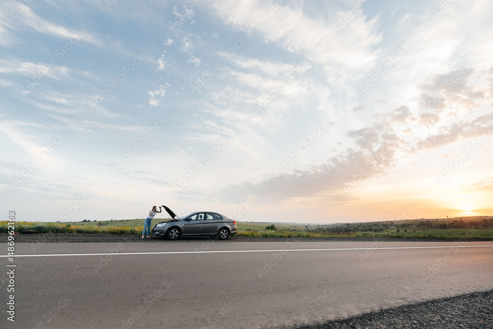 A young girl stands near a broken-down car in the middle of the highway during sunset and tries to call for help on the phone. Waiting for help. Car service. Car breakdown on road.