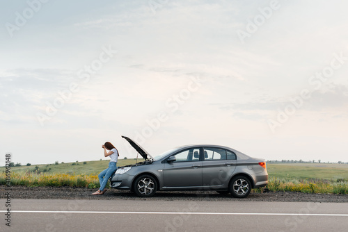 A young girl stands near a broken car in the middle of the highway during sunset and tries to call for help on the phone and start the car. Waiting for help. Car service. Car breakdown on the road. © Andrii