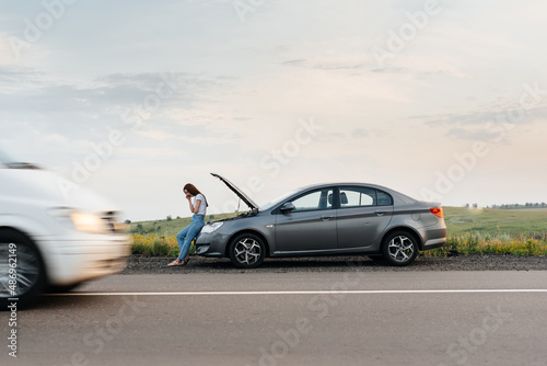 A young girl stands near a broken car in the middle of the highway during sunset and tries to call for help on the phone and start the car. Waiting for help. Car service. Car breakdown on the road. © Andrii