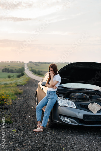 A frustrated young girl stands near a broken-down car in the middle of the highway during sunset. Breakdown and repair of the car. Waiting for help. Car service. Car breakdown on road. © Andrii