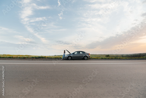 A young girl stands near a broken-down car in the middle of the highway during sunset and tries to call for help on the phone. Waiting for help. Car service. Car breakdown on road. © Andrii