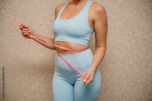 Cropped view of slim woman measuring waist with tape measure at home, close up. Unrecognizable european woman checking the result of diet for weight loss or liposuction indoors