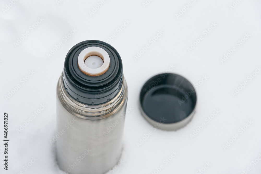 Thermos with a hot drink and a cup on a background of snow. Tourist utensils. Drink hot tea in winter to keep warm. Thermos with a hot drink and a cup on a background of snow. Tourist utensils. Drink hot tea in winter to keep warm.