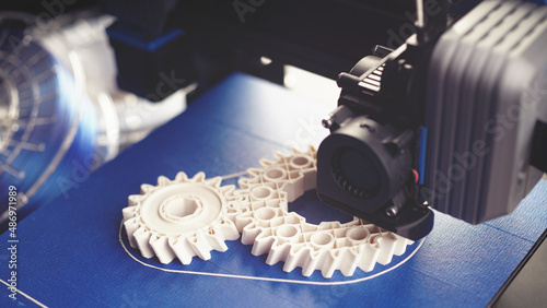 white helical gears produced by small black FDM-printer on blue build plate. clean bright technical sceney in wide format with filament rolls in background. excessive selective focus. 