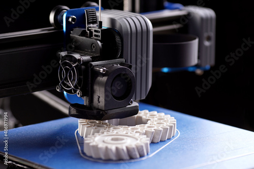 gantry with x-carriage and print head of a FDM-3D-printer that produces white helical gears on blue print bed. dark surrounding. selective focus. additive manufacturing concept photo