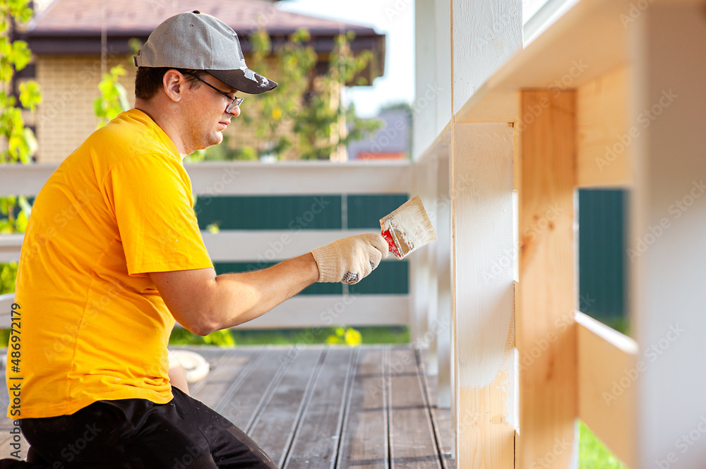 A man in a bright yellow T-shirt and cap holds a brush in his hand and paints the boards on the porch of the house with white paint A man in a bright yellow T-shirt and cap holds a brush in his hand and paints the boards on the porch of the house with white paint