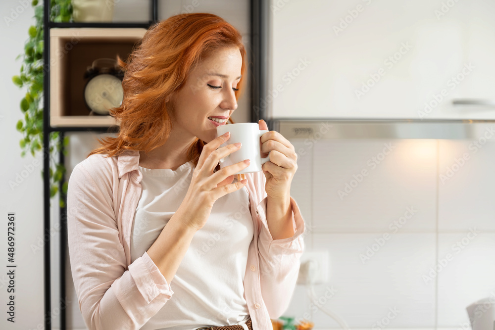 Closeup portrait of pretty red haired lady holding hot beverage in hands and enjoying amazing drink smell while standing in domestic kitchen at home Closeup portrait of pretty red haired lady holding hot beverage in hands and enjoying amazing drink smell while standing in domestic kitchen at home
